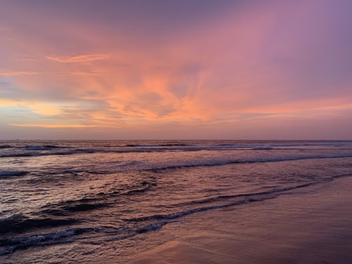A serene ocean view during sunset, with gentle waves lapping against the shore and the sky painted in warm hues of orange, pink, and purple. The horizon stretches far into the distance, creating a calm and tranquil atmosphere.
