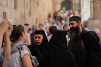 A group of people in traditional religious attire, including robes and head coverings, are gathered in an outdoor setting. The scene is bustling with a large crowd in the background, creating a sense of a busy street or public area. Stone walls and arched architecture suggest an old or historic environment.