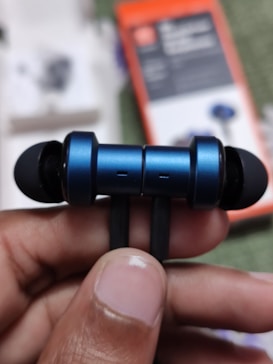 A close-up view of a hand holding a pair of blue wireless earphones. The focus is on the earbuds, which have a metallic blue finish with black tips. In the background, there is an out-of-focus box packaging.