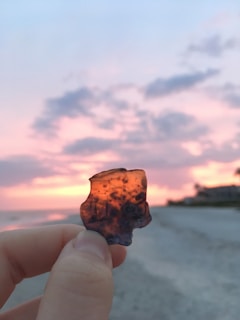 A hand holds a small piece of amber-colored translucent material against a backdrop of a vibrant sunset. The sky is painted with hues of pink, orange, and purple, with soft, blurred clouds. The horizon is visible in the background, possibly indicating a beach or coastal area.