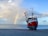 A red fishing boat is on a sandy beach near a calm sea. A vibrant rainbow arcs in the background beneath a partly cloudy sky. The setting provides a peaceful and serene atmosphere.
