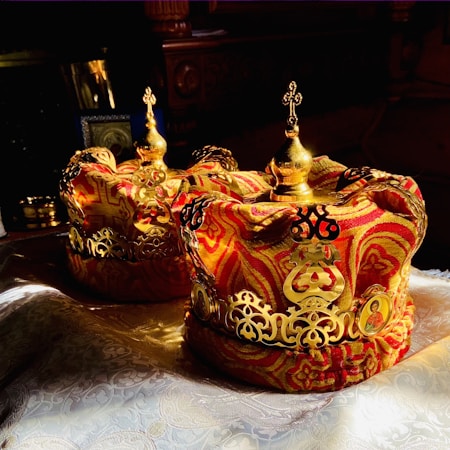 Luxurious crowns adorned with intricate gold embellishments and vibrant red and gold fabric resting on a richly textured surface. The crowns feature ornate patterns and religious symbols, suggesting ceremonial significance.