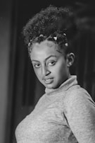 A grayscale portrait of a person with curly hair styled in an updo with small decorative elements. They are wearing a light-colored turtleneck sweater and are posed with a slight smile, looking directly at the camera.