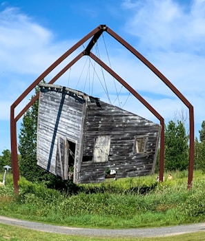 A weathered, rustic wooden cabin is suspended in mid-air by a large, geometric metal frame. The cabin appears aged, with peeling paint and an open door. It is surrounded by lush green grass and bushes under a clear blue sky.