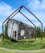 A weathered, rustic wooden cabin is suspended in mid-air by a large, geometric metal frame. The cabin appears aged, with peeling paint and an open door. It is surrounded by lush green grass and bushes under a clear blue sky.