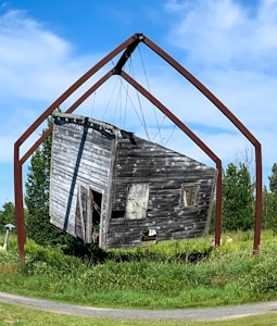 A weathered, rustic wooden cabin is suspended in mid-air by a large, geometric metal frame. The cabin appears aged, with peeling paint and an open door. It is surrounded by lush green grass and bushes under a clear blue sky.