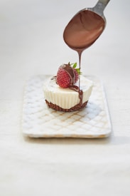 A small white dessert with a crumbly brown base is placed on a rectangular white plate with a quilted pattern. On top of the dessert is a fresh strawberry, and chocolate sauce is being poured over it from a spoon.