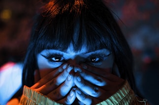 A close-up of a person with long eyelashes covering their lower face with their hands. The lighting casts a dramatic mix of blue and orange hues, highlighting the eyes and creating a mysterious atmosphere.