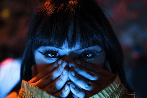A close-up of a person with long eyelashes covering their lower face with their hands. The lighting casts a dramatic mix of blue and orange hues, highlighting the eyes and creating a mysterious atmosphere.