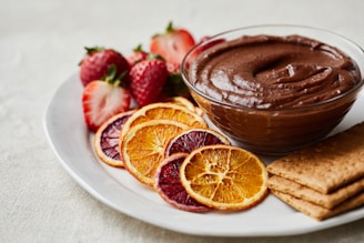 An appetizing display on a white plate includes a bowl of creamy chocolate dip, surrounded by sliced dried oranges, fresh strawberries, and a few rectangular graham crackers. The elements are arranged elegantly, suggesting a gourmet dessert or snack setup.