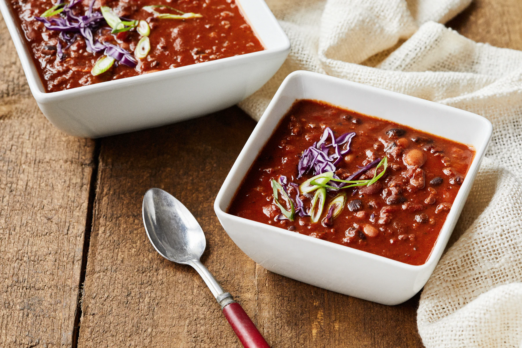 Two square white bowls of bean chili topped with scallions and red cabbage, a spoon beside them