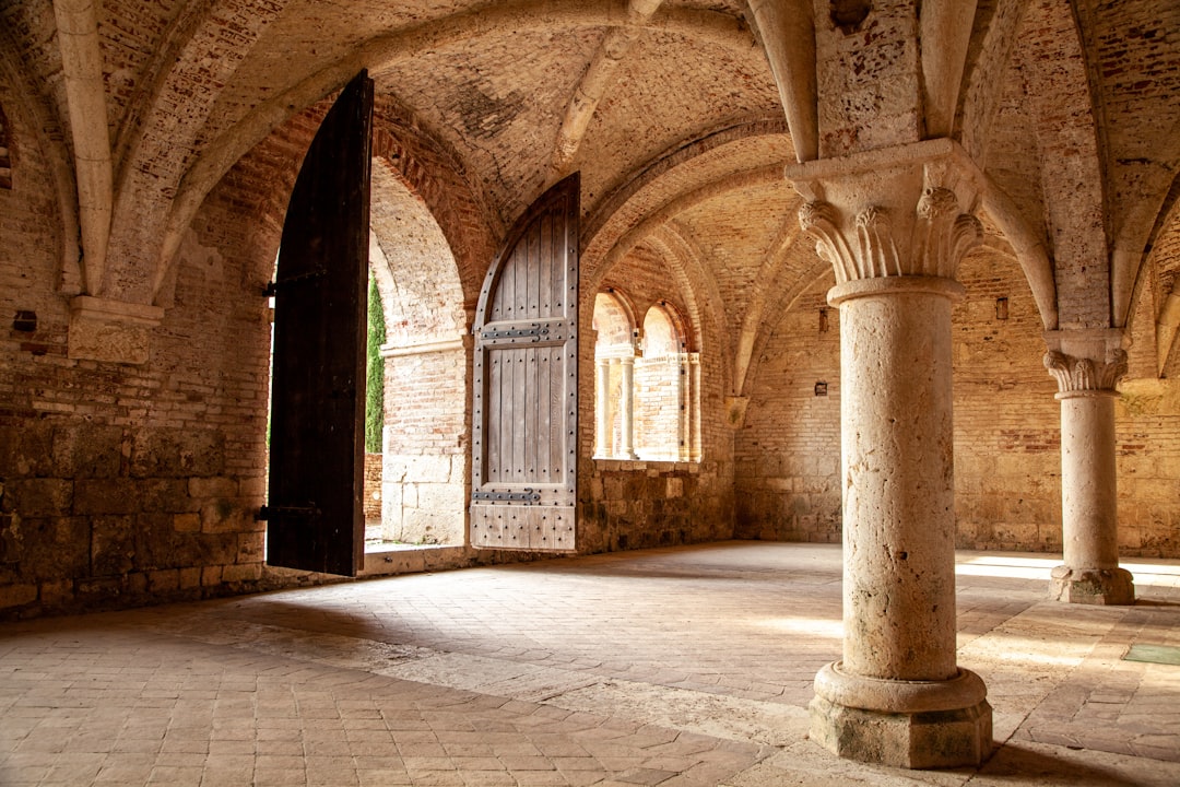 a large room with columns and a door, When visiting the San Galgano Abbey ruins in Tuscany, this vaulted building adjacent to the ruins of the abbey caught my attention as the sunset light created a warm effect, softening the shadows.</p>
<p></p>
<p>In this picture I also enjoy the "three-dimensional" effect of the various elements, displayed at an ever increasing distance from the camera lens. Worth also checking out the different texture of columns, floor tiles, ceiling vaults as well as the bolted wooden door.