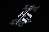 A large space station with multiple solar panels is floating against a backdrop of space. The station consists of several modular components and is symmetrical in design. The panels are arranged in a grid-like pattern, reflective of light.
