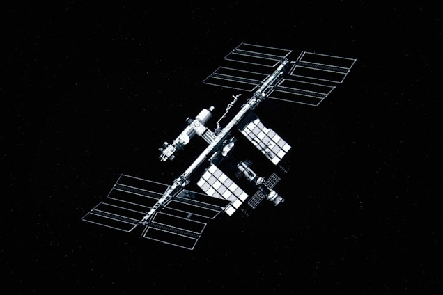 A large space station with multiple solar panels is floating against a backdrop of space. The station consists of several modular components and is symmetrical in design. The panels are arranged in a grid-like pattern, reflective of light.