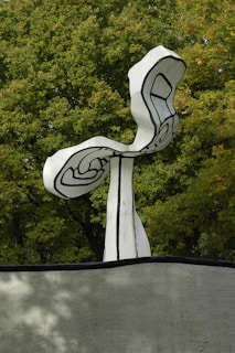 A black and white abstract sculpture with swirling patterns stands prominently against a backdrop of lush green trees. The sculpture's organic, curving form contrasts sharply with the angular lines that decorate its surface. It is situated on top of a grey concrete base.