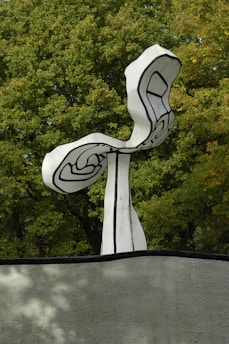 A black and white abstract sculpture with swirling patterns stands prominently against a backdrop of lush green trees. The sculpture's organic, curving form contrasts sharply with the angular lines that decorate its surface. It is situated on top of a grey concrete base.