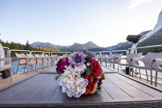 A vibrant bouquet of flowers sits on a table with a scenic backdrop of mountains and a lake. The arrangement includes a mix of colorful blooms such as red roses and purple dahlias. Surrounding the table, there are empty chairs arranged in a neat row, suggesting a setting prepared for an event or gathering.