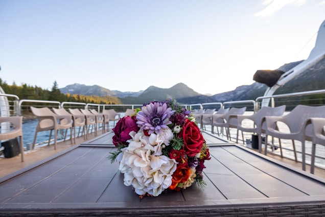 A vibrant bouquet of flowers sits on a table with a scenic backdrop of mountains and a lake. The arrangement includes a mix of colorful blooms such as red roses and purple dahlias. Surrounding the table, there are empty chairs arranged in a neat row, suggesting a setting prepared for an event or gathering.