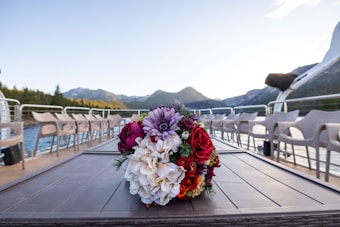 A vibrant bouquet of flowers sits on a table with a scenic backdrop of mountains and a lake. The arrangement includes a mix of colorful blooms such as red roses and purple dahlias. Surrounding the table, there are empty chairs arranged in a neat row, suggesting a setting prepared for an event or gathering.