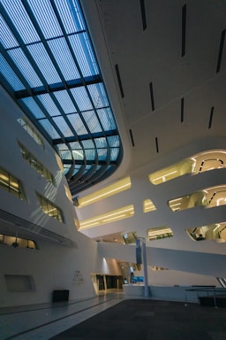 A modern architectural interior features sleek lines and geometric shapes, with large rectangular windows allowing natural light to stream in. The ceiling is designed with an intricate pattern of blue-tinted panels. The overall space is open and expansive, with a minimalist aesthetic.