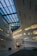 A modern architectural interior features sleek lines and geometric shapes, with large rectangular windows allowing natural light to stream in. The ceiling is designed with an intricate pattern of blue-tinted panels. The overall space is open and expansive, with a minimalist aesthetic.