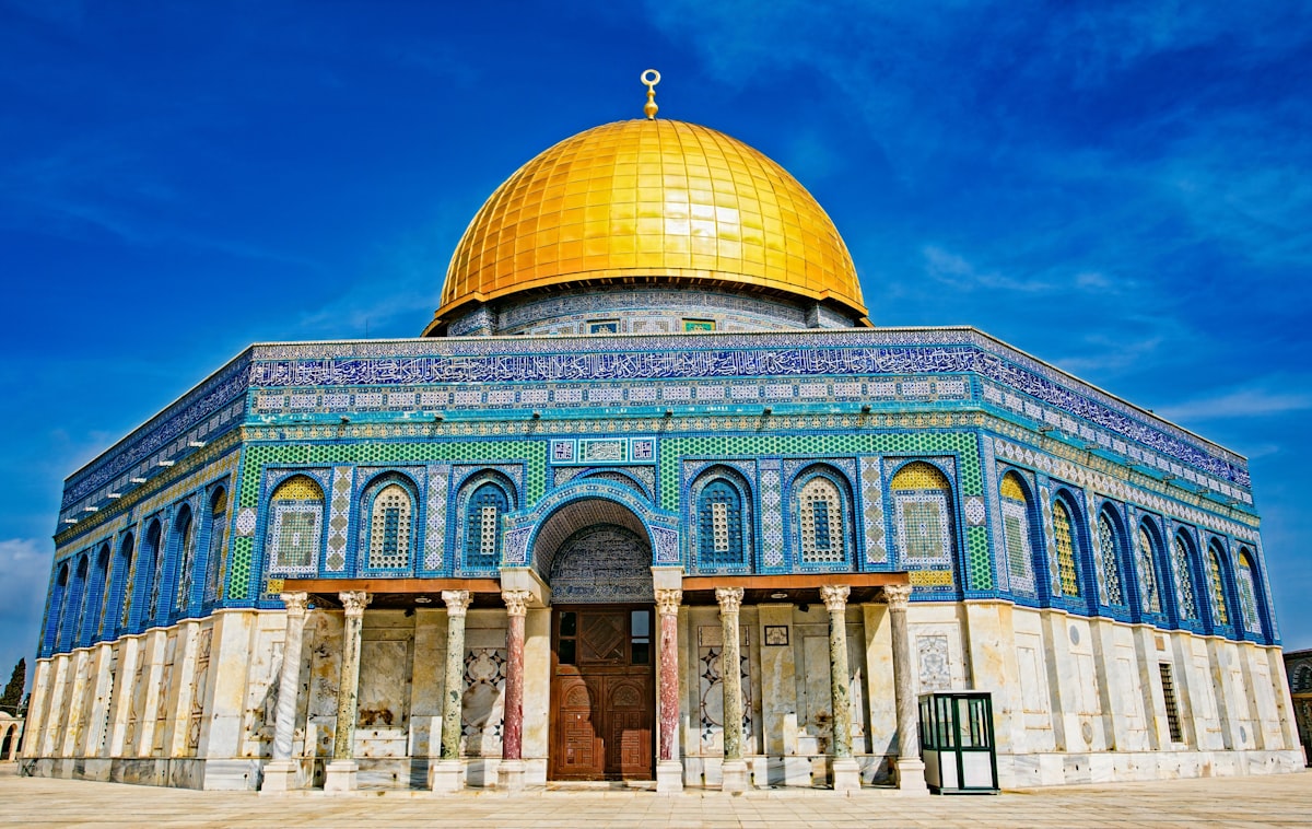 מבנה בעל כיפת זהב מבריקה הניצבת על גוף בנוי שצדדיו מצופים אריחים בכחול, טורקיז ולבן עם כתובות. בקומה התחתונה שורת עמודי שיש ומעליהם קשתות. השמיים כחולים עמוקים.