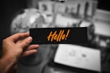 A hand holding a black card with the word 'Hello!' written in bold, orange letters. The background is blurred, showing a globe and some open notebooks or papers on a desk.