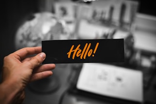 A hand holding a black card with the word 'Hello!' written in bold, orange letters. The background is blurred, showing a globe and some open notebooks or papers on a desk.