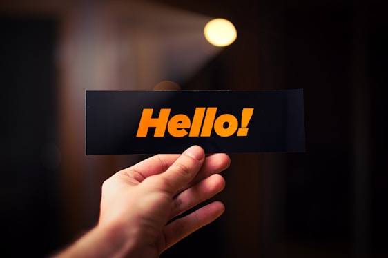 A hand holding a black card with the word 'Hello!' written in bold, orange letters. The background is out of focus with warm lighting, creating a cozy atmosphere.