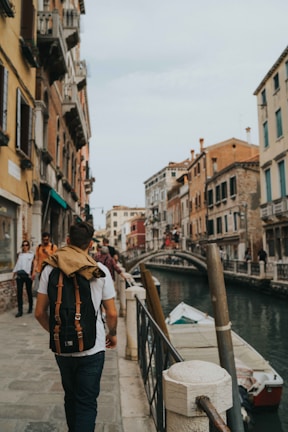 A man with a backpack walks along a canal in a historic city with narrow streets and colorful buildings. Others are also seen strolling along the path lined with a railing. A small boat is moored in the canal, and a stone bridge can be seen in the background connecting the two sides of the canal.