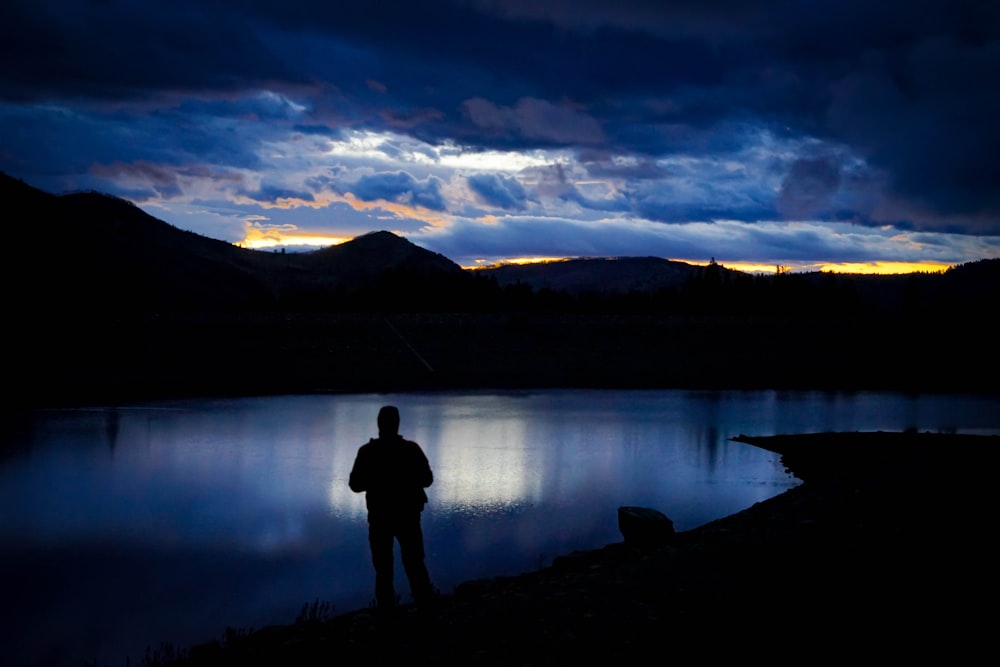 a man standing in front of a lake under a cloudy sky