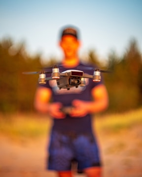 A drone is in focus, hovering in front of a blurred person who appears to be operating it. The setting is outdoors with natural lighting, possibly in a sandy or grassy area.