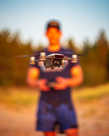 A drone is in focus, hovering in front of a blurred person who appears to be operating it. The setting is outdoors with natural lighting, possibly in a sandy or grassy area.