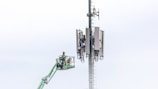 A worker in a safety harness is elevated on a green cherry picker, performing maintenance work on a large telecommunications tower equipped with multiple antennas against a light blue sky.