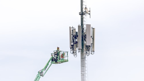 A worker in a safety harness is elevated on a green cherry picker, performing maintenance work on a large telecommunications tower equipped with multiple antennas against a light blue sky.