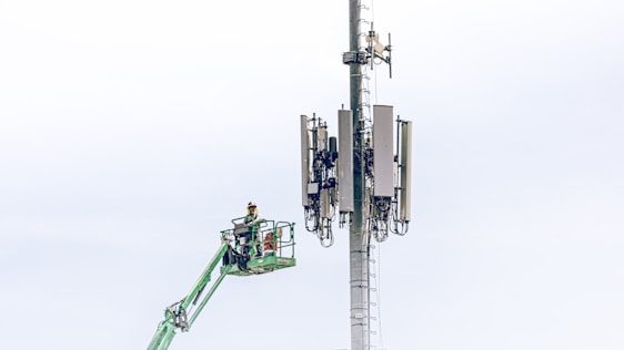 A worker in a safety harness is elevated on a green cherry picker, performing maintenance work on a large telecommunications tower equipped with multiple antennas against a light blue sky.