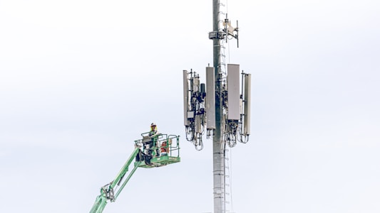 A worker in a safety harness is elevated on a green cherry picker, performing maintenance work on a large telecommunications tower equipped with multiple antennas against a light blue sky.