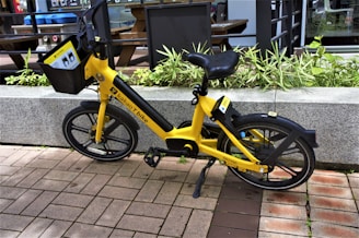 A bright yellow bicycle is parked on a brick pathway next to a concrete planter filled with green plants. The bike features a front basket displaying instructions and has a logo on its frame indicating it is a 'Kakao T bike.' The background includes a modern glass and metal building with outdoor furniture partly visible.