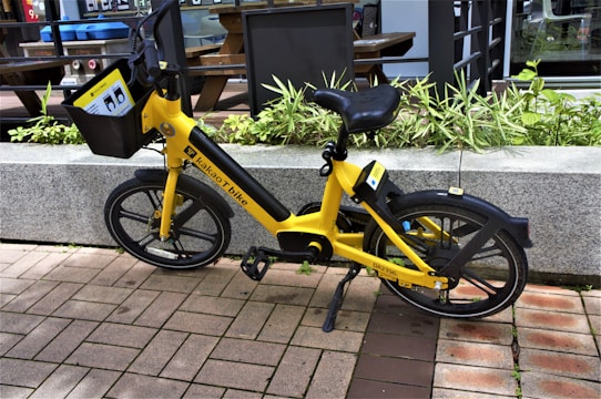 A bright yellow bicycle is parked on a brick pathway next to a concrete planter filled with green plants. The bike features a front basket displaying instructions and has a logo on its frame indicating it is a 'Kakao T bike.' The background includes a modern glass and metal building with outdoor furniture partly visible.