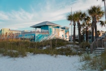 A vibrant blue building with a covered deck and white railings stands prominently along a sandy beach. The foreground features sea oats swaying in the breeze with a wooden walkway leading up to the structure. To the right, tall palm trees rise against a mostly clear sky, while American flags flutter in the distance.