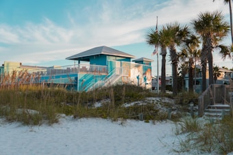 A vibrant blue building with a covered deck and white railings stands prominently along a sandy beach. The foreground features sea oats swaying in the breeze with a wooden walkway leading up to the structure. To the right, tall palm trees rise against a mostly clear sky, while American flags flutter in the distance.