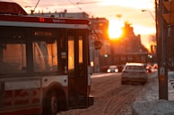 A city street scene during sunset with a red and white hybrid electric bus on the left side and several cars on the road. The sun is low in the sky, casting a warm, golden glow over the scene. Snow is visible on the edges of the road, and there is a pedestrian button on a nearby post.