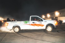 A white U-Haul pickup truck with the price $19.95 emblazoned on the side is parked on an asphalt surface, surrounded by a smoky atmosphere. Several people are visible around the truck, with some wearing hats and jackets. The background features an industrial-looking building, illuminated by bright lights, creating a night-time scene.