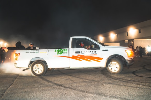 A white U-Haul pickup truck with the price $19.95 emblazoned on the side is parked on an asphalt surface, surrounded by a smoky atmosphere. Several people are visible around the truck, with some wearing hats and jackets. The background features an industrial-looking building, illuminated by bright lights, creating a night-time scene.