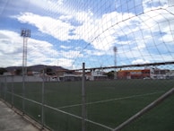 A fenced football field under a partly cloudy sky, surrounded by buildings and trees, with tall floodlights towering over the scene.