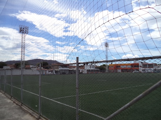 A fenced football field under a partly cloudy sky, surrounded by buildings and trees, with tall floodlights towering over the scene.
