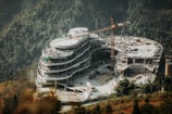 A large, modern building under construction surrounded by forested mountains. The structure is circular with multiple levels, and a crane towers above it. Construction materials and vehicles are visible on the ground around the site.