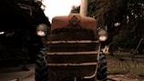 A rustic, vintage tractor with a weathered appearance, parked in an outdoor setting. The tractor features a prominent front grille and round headlights, with signs of age and rust. Trees and other farm equipment are visible in the background, creating a countryside atmosphere.