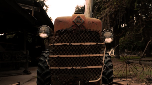 A rustic, vintage tractor with a weathered appearance, parked in an outdoor setting. The tractor features a prominent front grille and round headlights, with signs of age and rust. Trees and other farm equipment are visible in the background, creating a countryside atmosphere.