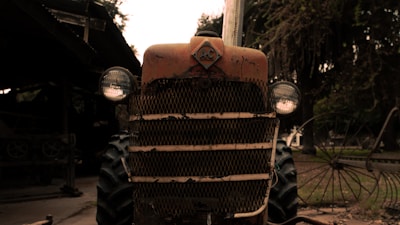 A rustic, vintage tractor with a weathered appearance, parked in an outdoor setting. The tractor features a prominent front grille and round headlights, with signs of age and rust. Trees and other farm equipment are visible in the background, creating a countryside atmosphere.